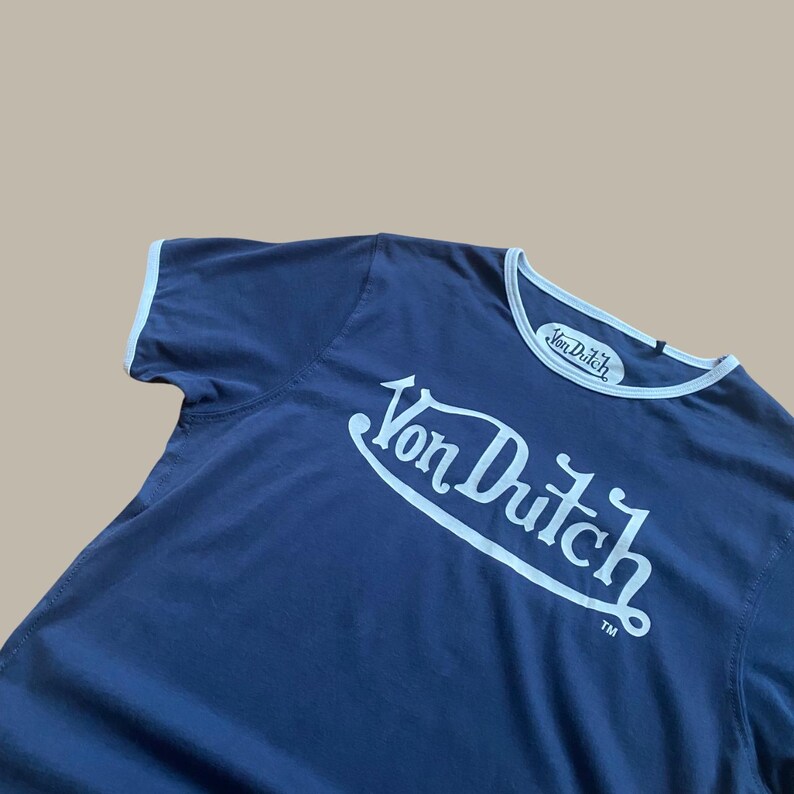 Puede incluir: Camiseta azul marino con ribetes blancos y el texto "Von Dutch" en una fuente estilizada. La camiseta tiene un estilo cl&aacute;sico de cuello redondo con cuello y bordes de manga blancos. El logotipo de Von Dutch es prominente.