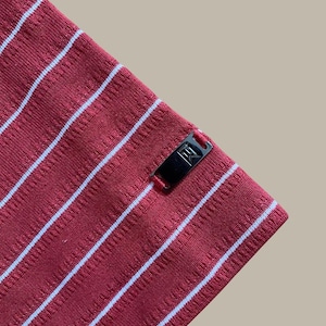 May include: Close-up of a red fabric with white stripes. The fabric has a textured appearance and a small black tag with white text. The stripes run diagonally across the image.