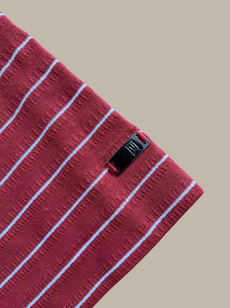 May include: Close-up of a red fabric with white stripes. The fabric has a textured appearance and a small black tag with white text. The stripes run diagonally across the image.