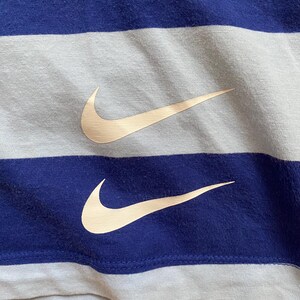 K&ouml;nnte beinhalten: Nahaufnahme eines blau-wei&szlig; gestreiften Stoffes mit zwei beigefarbenen Nike Swoosh-Logos. Der Stoff hat ein strukturiertes Aussehen, und die Logos sind auf den blauen Streifen positioniert.
