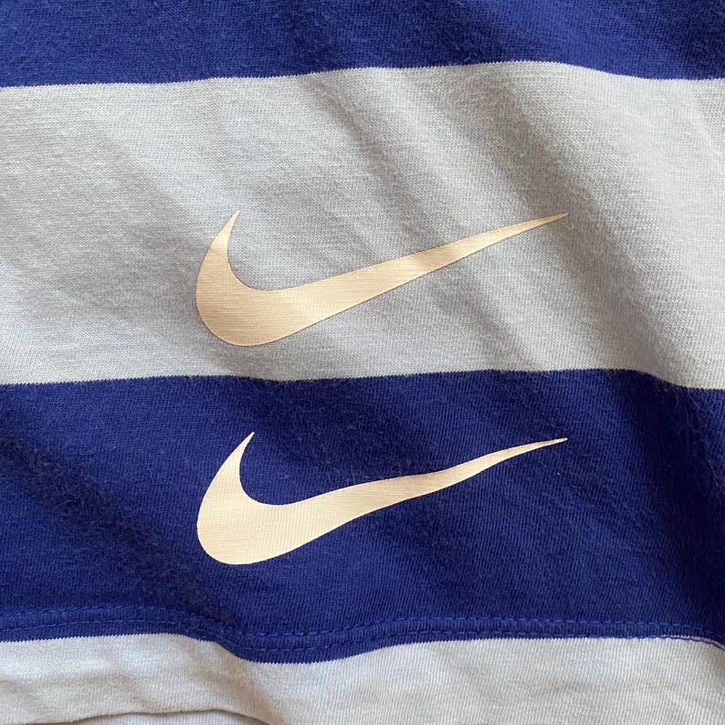 K&ouml;nnte beinhalten: Nahaufnahme eines blau-wei&szlig; gestreiften Stoffes mit zwei beigefarbenen Nike Swoosh-Logos. Der Stoff hat ein strukturiertes Aussehen, und die Logos sind auf den blauen Streifen positioniert.