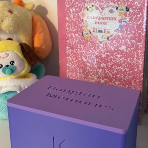 May include: A lilac box with the words "Bangtan Memories" embossed on the top. The box has a logo on the front and is next to a pink composition book and a plush toy.