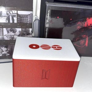 May include: A rectangular storage box with a red textured base and a white lid. The lid features three red circular and horizontal line symbols. The front of the red base displays a debossed BTS logo. The box is positioned in front of music memorabilia.