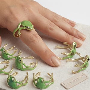 May include: A collection of gold-toned rings shaped like frogs with green enamel detailing on the back. Each ring features a frog with extended legs that wrap around the finger. The rings have small black eyes and a smooth, polished finish.