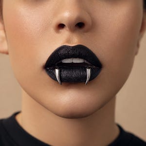 May include: A pair of silver-toned metal fangs worn as a lip piercing. The fangs are positioned on the lower lip, extending upwards over the matte black lipstick. The jewelry features a smooth, curved design that mimics the appearance of sharp teeth.