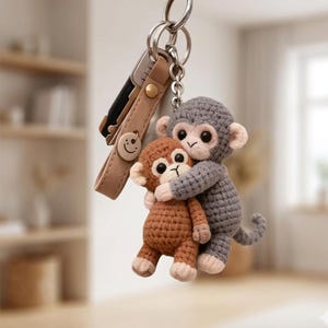 Op de afbeelding: Een handgemaakte sleutelhanger met twee gehaakte apenfiguren. Een grijze aap omhelst een bruine aap. De sleutelhanger bevat een leren bandje met een smiley-knop en een metalen sleutelring.