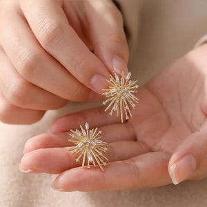 May include: A pair of gold-colored stud earrings with a starburst design. Each earring features multiple small, clear crystals and elongated, rectangular crystals radiating outwards. The earrings are held in a person's hand.