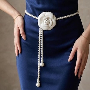 May include: A waist belt featuring a string of white faux pearls and a large, central white fabric camellia flower. Two long strands of pearls hang down from the flower, each finished with a single, larger white pearl at the end.