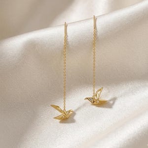 Peut inclure: Une paire de boucles d'oreilles pendantes dorées avec des motifs d'oiseaux en origami. Chaque boucle d'oreille est ornée d'une fine chaîne et d'un petit accent scintillant. Les boucles d'oreilles sont présentées sur un fond crème doux.