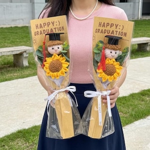 Peut inclure: Deux bouquets de remise de diplômes, chacun avec un tournesol crocheté et une petite figurine de diplômé portant un mortier. Les bouquets sont enveloppés de cellophane transparent et attachés avec des rubans blancs. Le texte "HAPPY:) GRADUATION" est imprimé en haut.