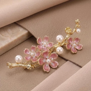 May include: A gold-toned brooch in the shape of a flowering branch. The brooch features pink enamel flowers with pearl centers and accents, along with several faux pearls. The design is delicate and elegant.