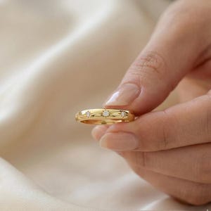 May include: A gold ring with three small, sparkling gemstones set in a row. The ring has a smooth, polished surface and a classic, elegant design. The ring is held in a hand against a soft, neutral background.
