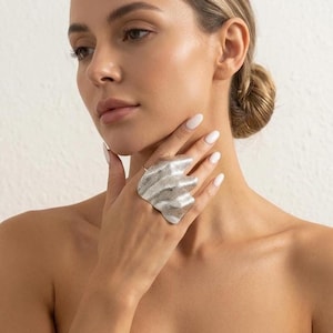 May include: A large, silver-toned statement ring featuring a wide, sculptural design with a textured, rippled surface that covers multiple fingers. The metal has a matte, hammered finish and an organic, wavy shape that extends across the hand.