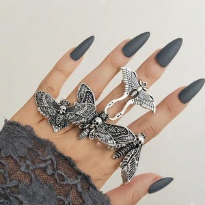 Könnte beinhalten: Ein silberfarbener Metallring mit drei Motiven von Nachtfaltern, die jeweils ein Totenkopfmuster auf dem Thorax aufweisen. Der Ring erstreckt sich über mehrere Finger und zeigt detaillierte Flügelstrukturen sowie ein poliertes Finish.