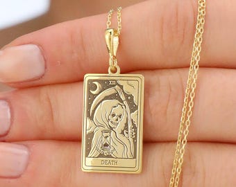 May include: Gold-colored pendant necklace featuring a rectangular charm with a detailed illustration of the Death tarot card. The charm depicts a hooded figure with a scythe, a crescent moon, and the word "DEATH".