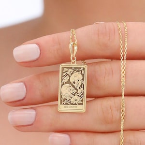 May include: A gold-colored pendant necklace featuring a rectangular charm with a detailed illustration of two figures and the text "THE LOVERS". The charm hangs from a delicate gold chain. The necklace is held up in front of a person.