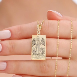 Może przedstawiać: Złoty naszyjnik z wisiorkiem w kształcie prostokąta z wygrawerowanym wizerunkiem uskrzydlonej postaci i napisem "TEMPERANCE". Naszyjnik jest trzymany przez osobę.