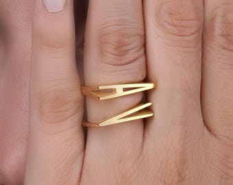 Könnte beinhalten: Ein goldfarbener Ring mit offenem Design, der die Buchstaben "A" und "V" zeigt. Der Ring wird an einem Finger getragen, wobei die Hand das Gesicht teilweise verdeckt. Der Ring hat einen modernen, minimalistischen Stil.