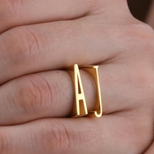 Puede incluir: Un anillo dorado con las letras "A" y "J" en un estilo de letras en bloque audaz. El anillo se lleva en un dedo, mostrando su diseño minimalista y su acabado pulido. El anillo es una joya personalizada.