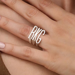 May include: A silver ring with the initials "RKG" in a stacked design. The ring is worn on a finger, with the initials standing out against the skin. The ring is a piece of jewelry.