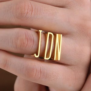 May include: A gold-colored ring with the letters "JDN" in a block letter style. The ring is worn on a finger, and the letters are raised and visible. The ring is a simple, yet elegant piece of jewelry.