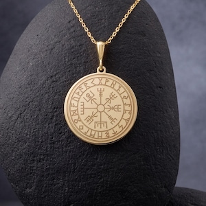 May include: A gold-colored pendant necklace featuring a circular pendant with engraved symbols. The pendant hangs from a delicate chain and is displayed on a black stone against a dark background. The pendant's design is intricate and detailed.