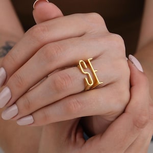 May include: A gold ring displaying the letters "SE" in a striking, modern font. The ring is worn on a finger, highlighting its design. The ring appears to be made of a shiny metal, reflecting light. The ring is a piece of jewellery.