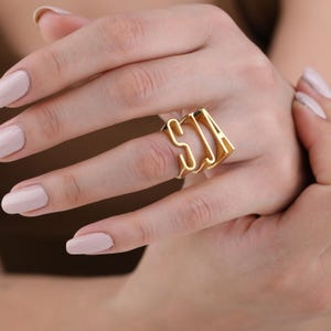 Può includere: Un anello color oro con le lettere "SJA" in un audace stile a blocchi. L'anello è indossato su un dito, mostrando il suo design personalizzato. La superficie lucida dell'anello riflette la luce, evidenziando la sua lucentezza metallica. Questo gioiello è un accessorio di tendenza.