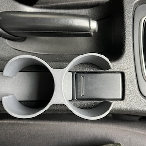 Op de afbeelding: Een grijze, 3D-geprinte bekerhouder voor in de auto met twee ronde openingen en een rechthoekig gedeelte. De bekerhouder is ontworpen om in de middenconsole van een auto te passen, met een zwarte handrem en versnellingspook op de achtergrond.