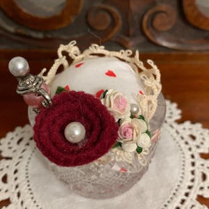 May include: A decorative pin cushion with a clear glass base, adorned with a burgundy felt flower, small pink and white paper roses, and a pearl accent. The cushion is topped with a white fabric dotted with red hearts.