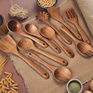 May include: A collection of wooden kitchen utensils, including spoons, spatulas, and slotted tools, arranged on a burlap surface. The utensils are made of light brown wood and vary in shape and size. Various ingredients are in small bowls in the background.