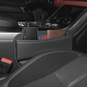 May include: Black car seat organizer with a pocket holding a smartphone, a credit card, and a brown wallet. The organizer is made of black leather and is positioned between the car seats. The car interior is black and gray.