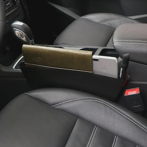 May include: Black car seat organizer with a wallet, smartphone, and pen. The organizer sits between the car seats and provides storage for small items. The wallet is olive green.