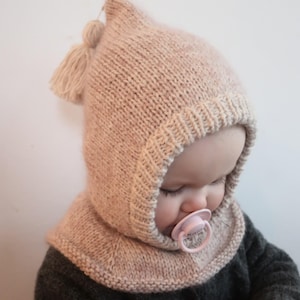 Può includere: Un cappello da bambino lavorato a maglia beige chiaro, con una parte superiore a punta, un piccolo nappa e un scaldacollo. Il cappello è realizzato con filato strutturato e copre la testa e il collo. Un ciuccio rosa è nella bocca del bambino.