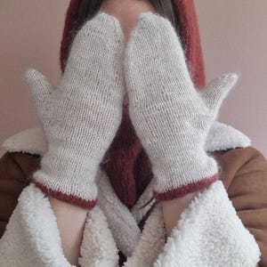 以下が含まれることがあります： オフホワイトのニットミトンで、袖口にダークレッドのトリムがあしらわれています。ミトンは、白いフェイクファーの襟と袖口が付いた茶色のジャケットの上に着用されており、頭部にはダークレッドのニットフードを被っています。
