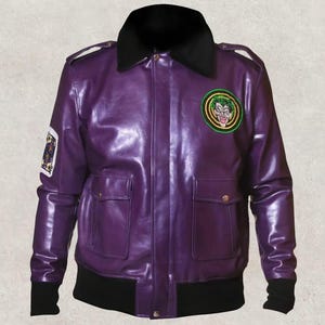 Può includere: Giacca in pelle viola con colletto e polsini neri. La giacca presenta due tasche frontali, spalline e una toppa circolare con un disegno Joker verde e giallo. Una toppa con carta da gioco è sulla manica sinistra.