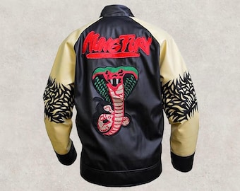 May include: Black satin jacket with a red and green cobra patch and the word "KING TUK" embroidered on the back. The sleeves are tan with black decorative patterns. The jacket has a black collar and cuffs.