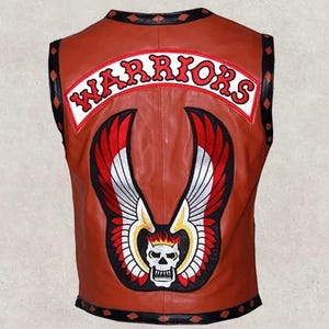Könnte beinhalten: Braune Lederweste mit großem, gesticktem Aufnäher auf dem Rücken. Der Aufnäher zeigt das Wort "WARRIORS" in roten Buchstaben, einen Totenkopf und ein geflügeltes Design in Rot, Weiß und Schwarz.