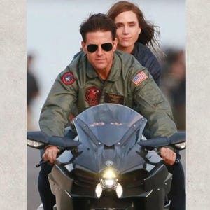 Könnte beinhalten: Ein Mann mit Sonnenbrille und grüner Militärjacke fährt ein schwarzes Motorrad, mit einer Frau als Beifahrerin. Das Motorrad hat eine dunkle Windschutzscheibe und Seitenspiegel. Der Hintergrund ist unscharf.