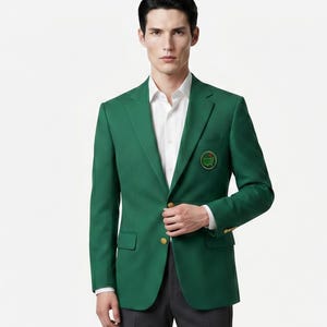 Puede incluir: Un blazer verde oscuro con un emblema dorado en el bolsillo del pecho. El blazer tiene botones dorados y se lleva sobre una camisa blanca. La persona lleva pantalones grises oscuros.