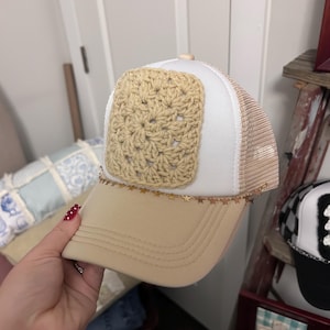 May include: A beige and white trucker hat featuring a square beige crochet patch on the front panel. The hat includes a beige brim and a mesh back. A delicate gold-toned chain with small star-shaped charms is attached across the base of the crown.