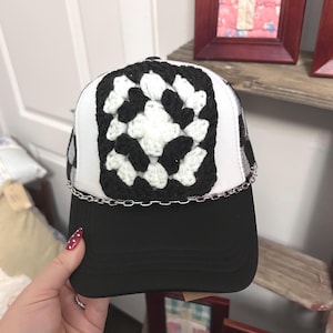 May include: A black and white baseball cap featuring a square crochet granny square patch on the front panel. The cap has a solid black brim and a silver-toned metal chain detail running horizontally across the base of the front panel.