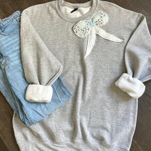 May include: A gray sweatshirt with a decorative bow made of floral and gingham fabric. The cuffs are white. Light blue jeans are folded next to the sweatshirt. The items are displayed on a wooden surface.