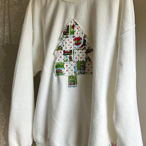 Könnte beinhalten: Weißes Sweatshirt mit einem Weihnachtsbaum-Design. Der Baum besteht aus Patchwork-Stoffen mit roten, grünen und weißen Akzenten. Das Sweatshirt hat einen Rundhalsausschnitt und lange Ärmel. Ein kleiner Aufnäher befindet sich am Ärmel.