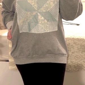 May include: A gray sweatshirt with a light blue and white patchwork design on the front. The patchwork features a pinwheel pattern with floral and gingham fabrics. The sweatshirt has a crew neck and long sleeves with white cuffs.