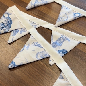 Può includere: Ghirlanda di bandierine in tessuto con un motivo di gallo e fiori blu e bianco. Le bandierine triangolari sono collegate da un nastro bianco, perfette per decorare una stanza o un evento. Le bandierine sono in tessuto.