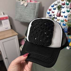 May include: A black and white trucker hat featuring a square, black crocheted granny square patch centered on the white foam front panel. The hat has a black brim, black mesh back panels, and a black adjustable snapback closure at the rear.