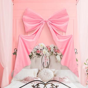 May include: A pink canopy bed with a large bow-shaped fabric decoration. The bed features a white headboard adorned with floral arrangements and decorative pillows. The overall aesthetic is romantic and feminine, with soft pink hues.