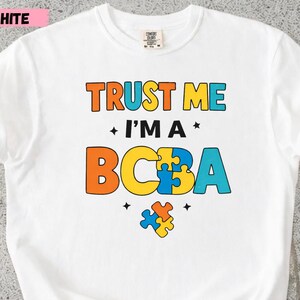 Könnte beinhalten: Weißes T-Shirt mit dem Text "TRUST ME I'M A BCBA" in Orange, Blau und Gelb. Das Design enthält Puzzleteile. Das Shirt ist aus einem weichen Material.