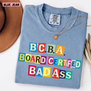 Puede incluir: Una camiseta azul claro con el texto "BCBA BOARD CERTIFIED BADASS" en letras de bloque coloridas. Un collar dorado con un colgante descansa sobre la camiseta. La camiseta está doblada sobre una superficie blanca.